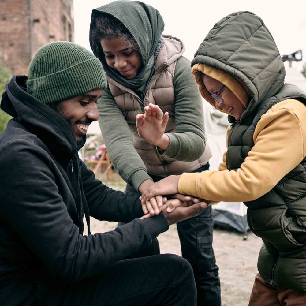 Positive young Black man in hat playing with kids against abandoned building and hanging clothes on rope in refugee camp